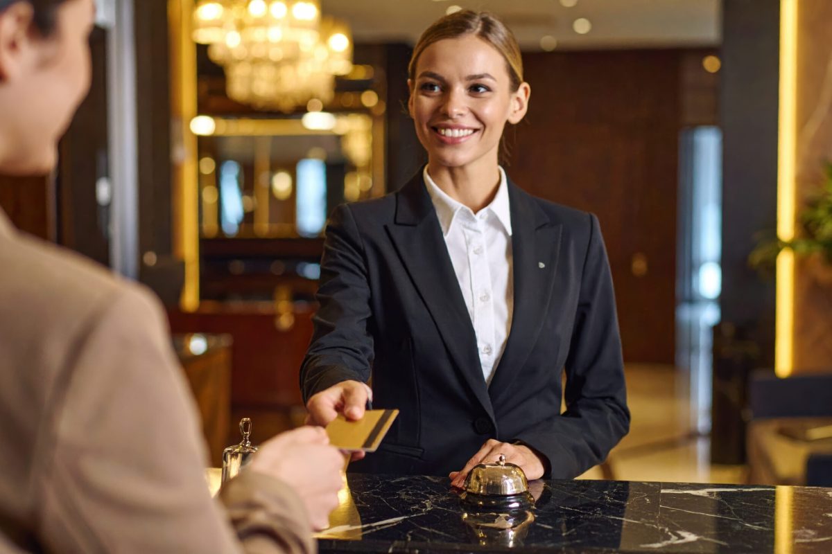 Smiling hotel receptionist in black suit accepts credit card from guest at front desk, with service bell and elegant interior lighting creating welcoming atmosphere
